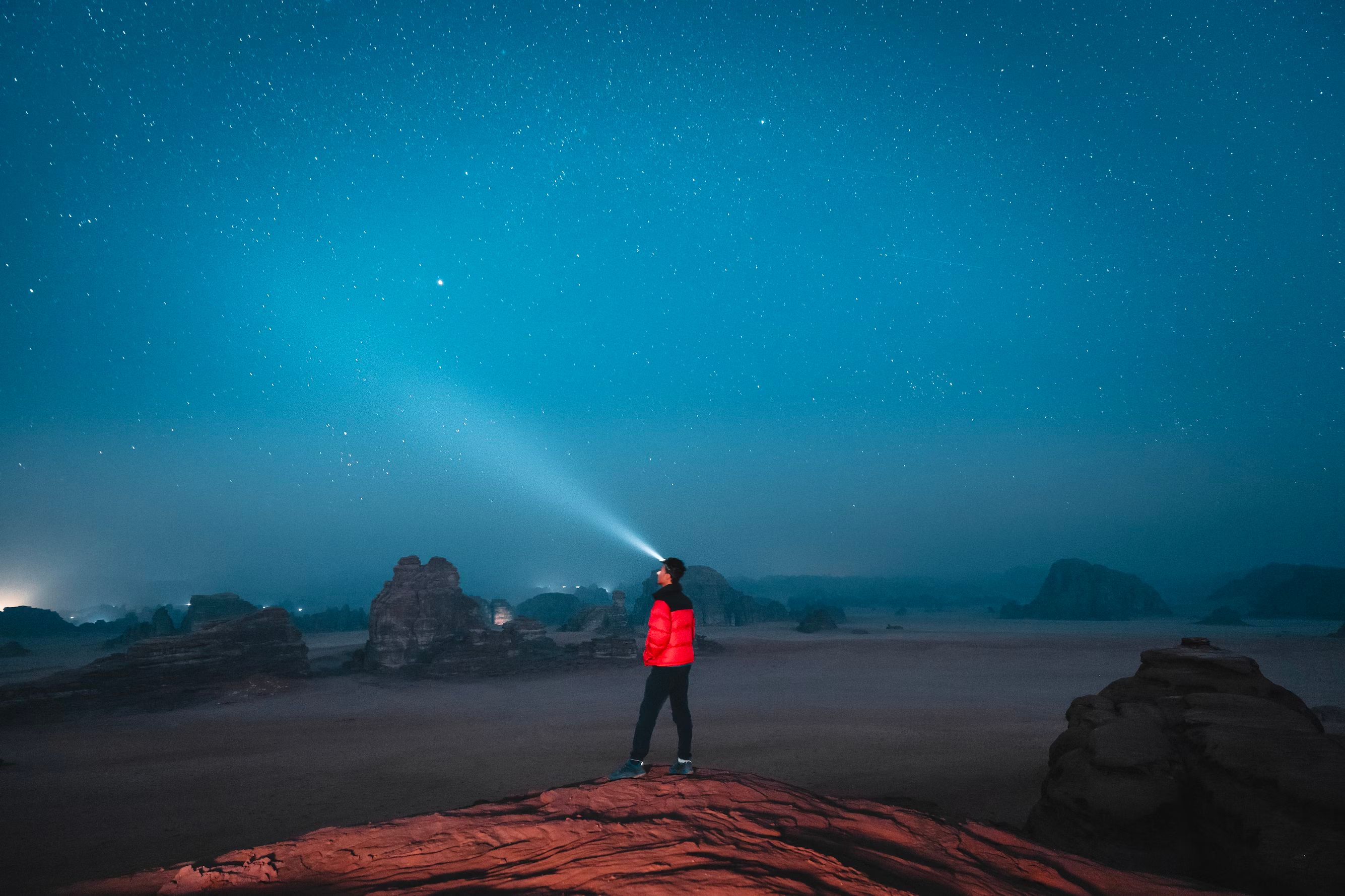 Hiker with headlamp viewing a vibrant night sky atop a rock formation in a place that looks like a national park